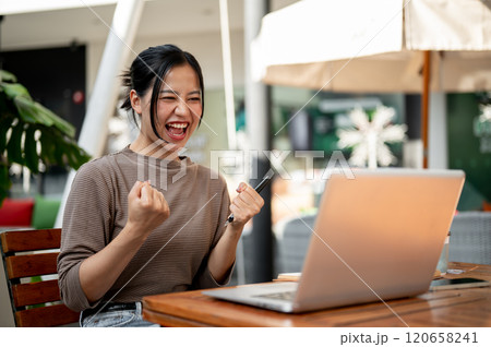 A woman sits in front of her laptop outdoors, celebrating her success with an excited scream of joy. A woman sits in front of her laptop outdoors, celebrating her success with an excited scream of joy. 120658241