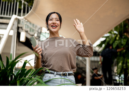 A cheerful Asian woman smiles brightly and waves as she unexpectedly runs into a friend outdoors. 120658253