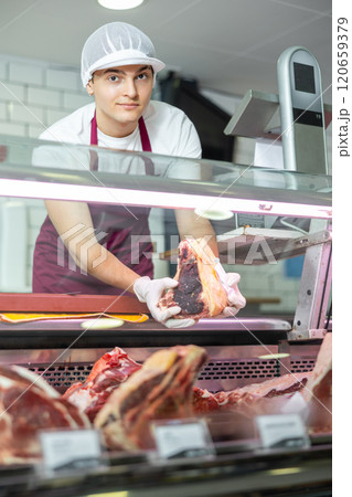 Positive young salesman demonstrating piece of meat in butcher shop Positive young salesman demonstrating piece of meat in butcher shop 120659379
