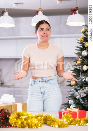 Upset young woman in casual clothes near wrapped gift and Christmas tree in living room at home 120659395