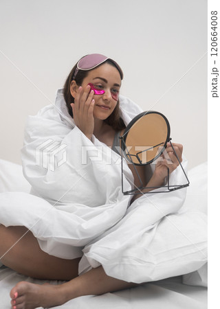 Cocooned in her blanket, a woman applies eye patches while seated on her bedroom bed, meticulously inspecting her face with a hand mirror.  120664008
