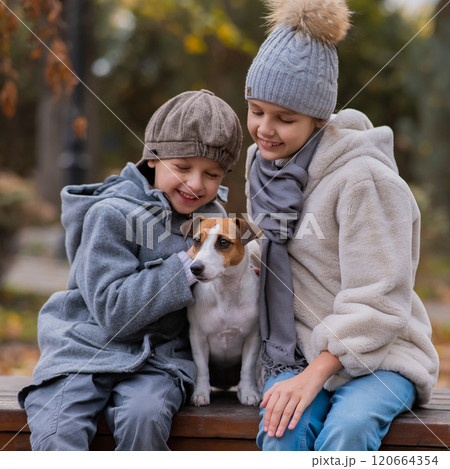 Brother and sister sit in an embrace with a dog on a bench for a walk in the autumn park. Boy, girl and jack russell terrier.  120664354