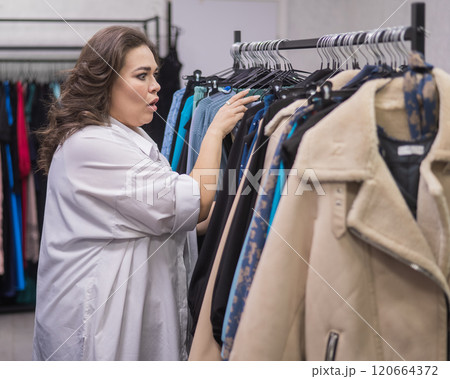 A smiling fat woman in a plus size store chooses clothes while sorting through the hangers.  120664372