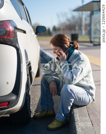 A Caucasian woman sits by a car wheel and calls for help on the phone. 120664429