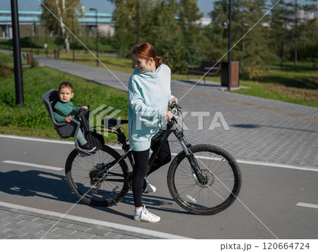 Caucasian woman riding bicycle with toddler son sitting behind her in child seat in park.  120664724