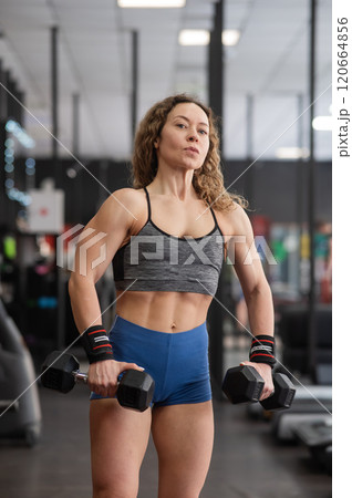 Muscular Caucasian forty year old woman doing exercises with dumbbells in the gym. Vertical photo. Muscular Caucasian forty year old woman doing exercises with dumbbells in the gym. Vertical photo. 120664856
