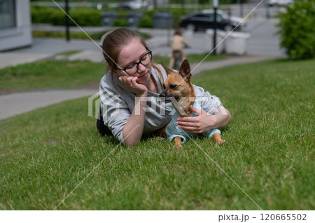 Young Caucasian woman lying on the lawn while walking with a small dog. Young Caucasian woman lying on the lawn while walking with a small dog. 120665502