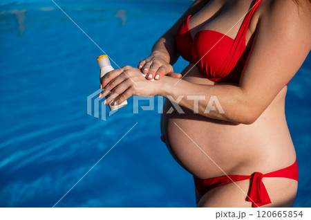 Close-up of the belly of a pregnant woman sunbathing in a red bikini. Expectant mother applying sunscreen to her hands. Close-up of the belly of a pregnant woman sunbathing in a red bikini. Expectant mother applying sunscreen to her hands. 120665854
