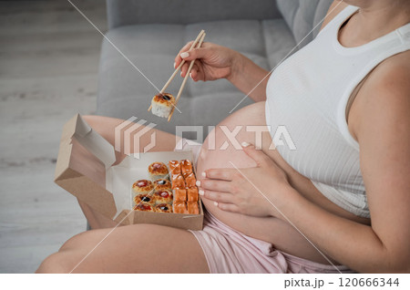 A pregnant woman sits on the sofa and eats rolls from a box. Food delivery. Close up of belly. A pregnant woman sits on the sofa and eats rolls from a box. Food delivery. Close up of belly. 120666344