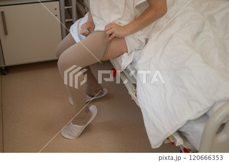 A woman puts on post-operative compression stockings in a hospital room. A woman puts on post-operative compression stockings in a hospital room. 120666353