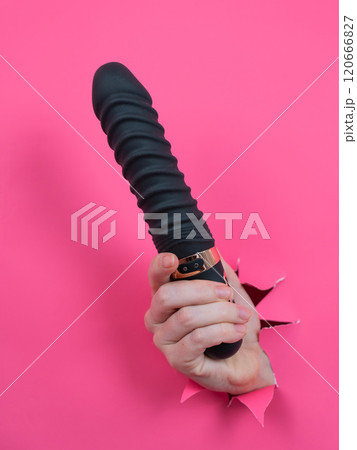 Female hand sticking out of pink paper background with black textured vibrator. Vertical photo. Female hand sticking out of pink paper background with black textured vibrator. Vertical photo. 120666827
