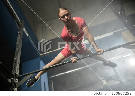 Low angle shot of professional sportswoman in straddle planche on high bar perfectly performing static hold while exercising calisthenics in gym with low key lighting, copy space 120667258
