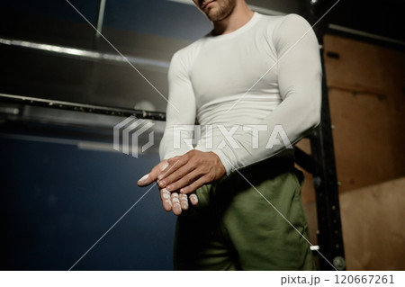 Cropped shot of professional sportsman dusting hands with chalk applying magnesia powder on palms for stronger grip on horizontal bar or barbell in gym with low key lighting, copy space 120667261