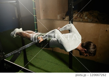 Strong sportsman doing back lever pull ups holding body straight while working out on high bar in gym with low key lighting, copy space Strong sportsman doing back lever pull ups holding body straight while working out on high bar in gym with low key lighting, copy space 120667262