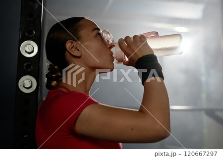Side view of fit young sportswoman hydrating with athlete drink after draining workout captured against sunbeam in gym with low key lightning, copy space 120667297