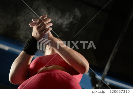 Low angle shot of unrecognizable female gymnast applying magnesia powder on palms clapping hands in chalk cloud in low key lit gym, copy space 120667310