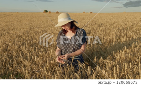 Beautiful Woman Stands In Wheat Field Wearing Dress And Hat Touches Spikelets With Her Hands. Freedom Concept. Enjoying Warm Evening. Moves slowly. Beautiful Woman Stands In Wheat Field Wearing Dress And Hat Touches Spikelets With Her Hands. Freedom Concept. Enjoying Warm Evening. Moves slowly. 120668675