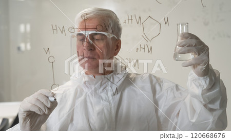 An elderly Caucasian male scientist in a protective suit holds a test tube and writes a formula on the glass.  120668676