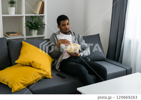 Cheerful curly haired ethnic african american male in casual clothes resting at home on comfortable sofa while watching tv channels or movie during weekend. Tv set, online television and entertiment Cheerful curly haired ethnic african american male in casual clothes resting at home on comfortable sofa while watching tv channels or movie during weekend. Tv set, online television and entertiment 120669263