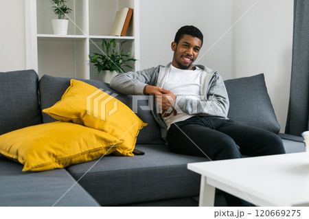 Cheerful curly haired ethnic african american male in casual clothes resting at home on comfortable sofa while watching tv channels or movie during weekend. Tv set, online television and entertiment Cheerful curly haired ethnic african american male in casual clothes resting at home on comfortable sofa while watching tv channels or movie during weekend. Tv set, online television and entertiment 120669275