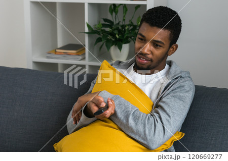 Cheerful curly haired ethnic african american male in casual clothes resting at home on comfortable sofa while watching tv channels or movie during weekend. Tv set, online television and entertiment Cheerful curly haired ethnic african american male in casual clothes resting at home on comfortable sofa while watching tv channels or movie during weekend. Tv set, online television and entertiment 120669277
