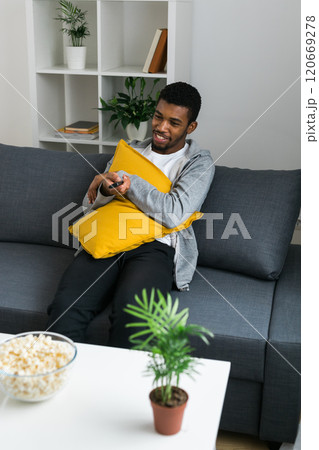 Cheerful curly haired ethnic african american male in casual clothes resting at home on comfortable sofa while watching tv channels or movie during weekend. Tv set, online television and entertiment Cheerful curly haired ethnic african american male in casual clothes resting at home on comfortable sofa while watching tv channels or movie during weekend. Tv set, online television and entertiment 120669278