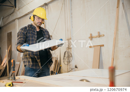 Carpenter man holding piece of wood surrounded by other wood pieces, showcasing craftsmanship and happiness at work in a furniture factory. Ideal for woodworking and craftsmanship concepts. Labor day Carpenter man holding piece of wood surrounded by other wood pieces, showcasing craftsmanship and happiness at work in a furniture factory. Ideal for woodworking and craftsmanship concepts. Labor day 120670974