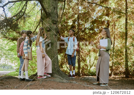 Group of teen students touching trunk of old tree during tour in botanical garden on warm autumn day, wide shot 120672012