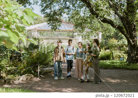 Wide shot of young woman working in botanical garden conducting tour for group of teenagers on sunny day 120672021
