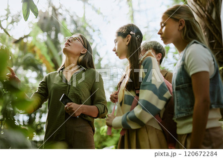 Low angle view of teen students visiting botanical garden, listening to female specialist speaking about plants growing in greenhouse Low angle view of teen students visiting botanical garden, listening to female specialist speaking about plants growing in greenhouse 120672284