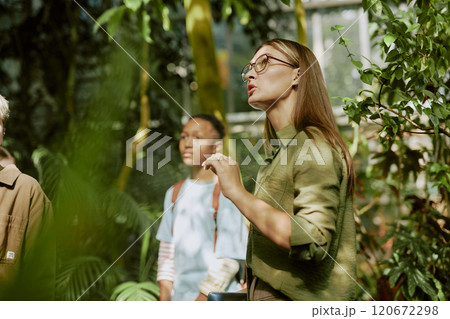 Young female tour guide speaking about rare plants growing in greenhouse while working with group of teenagers 120672298