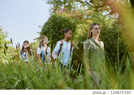 Young Caucasian woman walking along botanical garden, showing it to group of diverse teenagers, copy space 120672330
