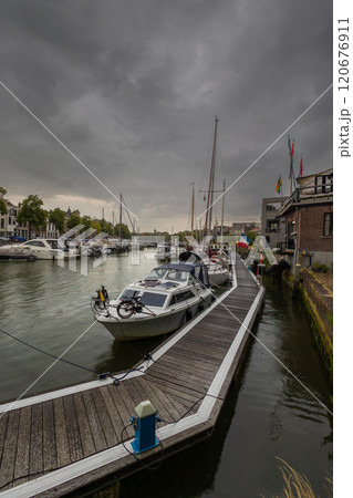 Serene harbor with moored boats under a dramatic cloudy sky. Quaint historic buildings and trees line the waterfront, reflecting in the calm green water, creating peaceful scene. Dordrecht landscape. 120676911