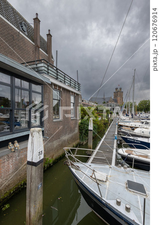Serene harbor with moored boats under a dramatic cloudy sky. Quaint historic buildings and trees line the waterfront, reflecting in the calm green water, creating peaceful scene. Dordrecht landscape. Serene harbor with moored boats under a dramatic cloudy sky. Quaint historic buildings and trees line the waterfront, reflecting in the calm green water, creating peaceful scene. Dordrecht landscape. 120676914