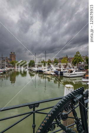 Serene harbor with moored boats under a dramatic cloudy sky. Quaint historic buildings and trees line the waterfront, reflecting in the calm green water, creating peaceful scene. Dordrecht landscape. Serene harbor with moored boats under a dramatic cloudy sky. Quaint historic buildings and trees line the waterfront, reflecting in the calm green water, creating peaceful scene. Dordrecht landscape. 120676915