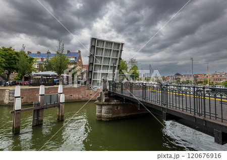 A canal scene with a historic drawbridge, lined with classic Dutch buildings and trees. Boats are docked under the cloudy sky, and a church tower is visible in the background. Dordrecht landscape. A canal scene with a historic drawbridge, lined with classic Dutch buildings and trees. Boats are docked under the cloudy sky, and a church tower is visible in the background. Dordrecht landscape. 120676916