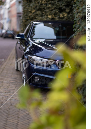 A sleek dark blue car parked on a cobblestone street, partially hidden behind greenery. The scene captures a blend of urban and natural elements with a touch of elegance. A sleek dark blue car parked on a cobblestone street, partially hidden behind greenery. The scene captures a blend of urban and natural elements with a touch of elegance. 120676919