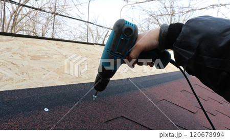 a hand in warm work clothes with a blue electric screwdriver screws fasteners into the roof structure of a low building, connecting soft tiles and an OSB sheet 120678994