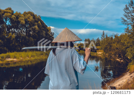 A female samurai stands by a river, holding a katana and looking into the distance on a sunny day. A samurai with a katana on his shoulder contemplates while looking at the calm surface of the river. A female samurai stands by a river, holding a katana and looking into the distance on a sunny day. A samurai with a katana on his shoulder contemplates while looking at the calm surface of the river. 120681173