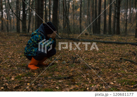 Sad child squatting in forest covering face crying lost during hike wearing orange rubber boots bright jacket and black hat. Scared child lost in forest crying. 120681177