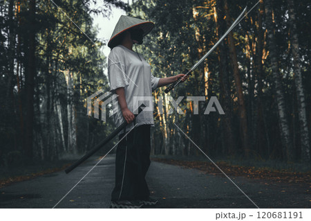 A warrior wearing a conical Asian hat and holding a katana stands on a paved path through the forest. The man's face is hidden by a black mask, and the katana is raised and ready for battle. A warrior wearing a conical Asian hat and holding a katana stands on a paved path through the forest. The man's face is hidden by a black mask, and the katana is raised and ready for battle. 120681191