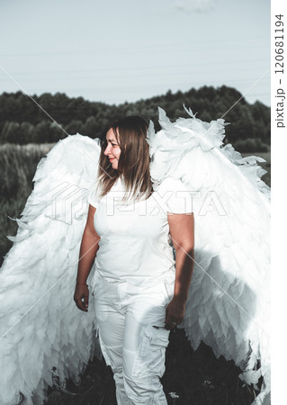 A woman with white angel wings stands in a field with trees and a blue sky in the background, creating a serene and ethereal atmosphere. Beautiful woman as a white angel. 120681194