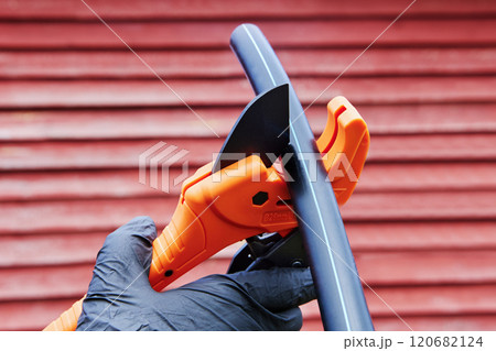 Plumber cuts HDPE pipe with pipe cutter with ratchet, when installing pipeline for drinking and irrigation water, close-up against background of red weathered wooden wall. 120682124