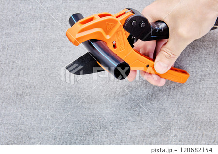 Hand of plumber holds pipeline scissors cutter with ratchet mechanism ready to cut segment of HDPE pipe, closeup on gray background. Hand of plumber holds pipeline scissors cutter with ratchet mechanism ready to cut segment of HDPE pipe, closeup on gray background. 120682154