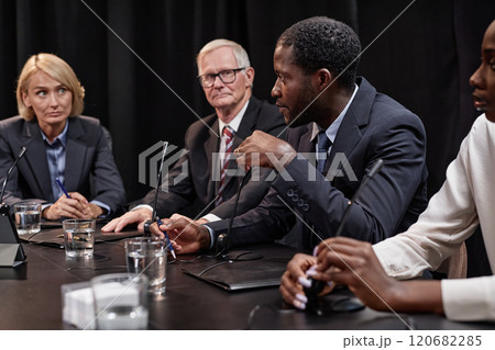 Side view of confident African American politician speaking into microphone debating with female colleague about initiative while sitting in line with other officials at meeting table, copy space 120682285