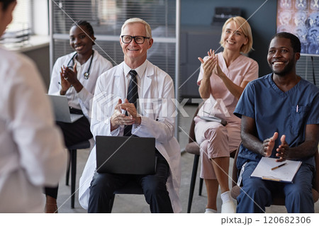 Smiling senior male head physician supporting speaker with applause sitting in conference room with diverse team of doctors during colleagues presentation Smiling senior male head physician supporting speaker with applause sitting in conference room with diverse team of doctors during colleagues presentation 120682306