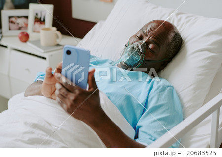 Elderly man lying in hospital bed holding smartphone, connected to oxygen, appearing calm and focus, medically equipped room surrounding him, showing caring environment 120683525