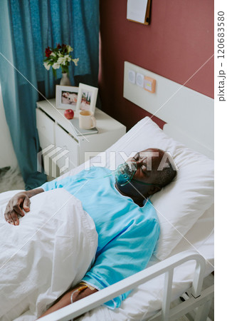 Elderly man resting in hospital bed, attached to medical equipment and storage visible beside bed including flowers and framed photo Elderly man resting in hospital bed, attached to medical equipment and storage visible beside bed including flowers and framed photo 120683580