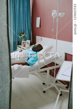 Resting African American patient in hospital bed, wearing oxygen mask, with clipboard and flowers on bedside table, signifying recovery process in healthcare environment 120683583