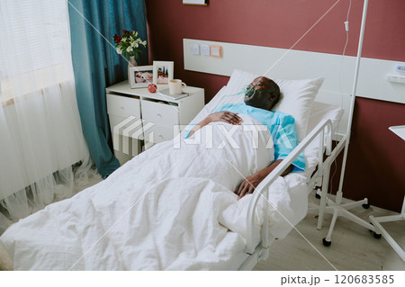 Hospital patient lying in bed beside window with light flowing in, surrounded by medical equipment and furnishings, wearing face mask for breathing assistance 120683585